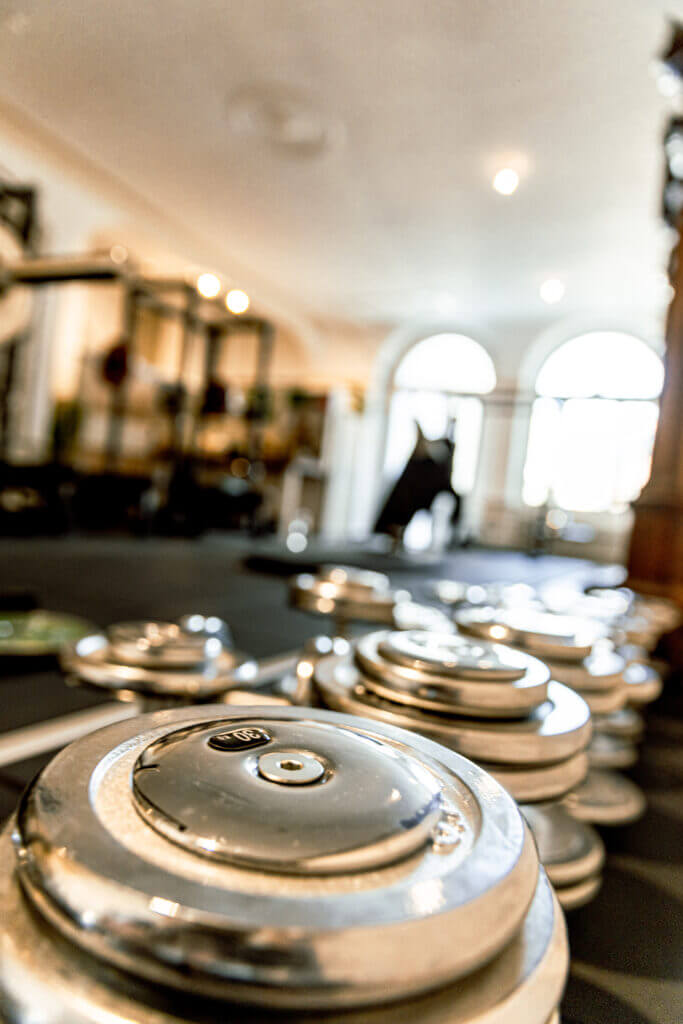 Nahaufnahme von silbern glänzenden Hantelscheiben, die auf dem Boden eines Fitnessstudios gestapelt sind, mit verschwommenen Fitnessgeräten und großen Fenstern im Hintergrund, die eine helle und konzentrierte Trainingsatmosphäre schaffen - ideal für Personal Training in Lübeck.