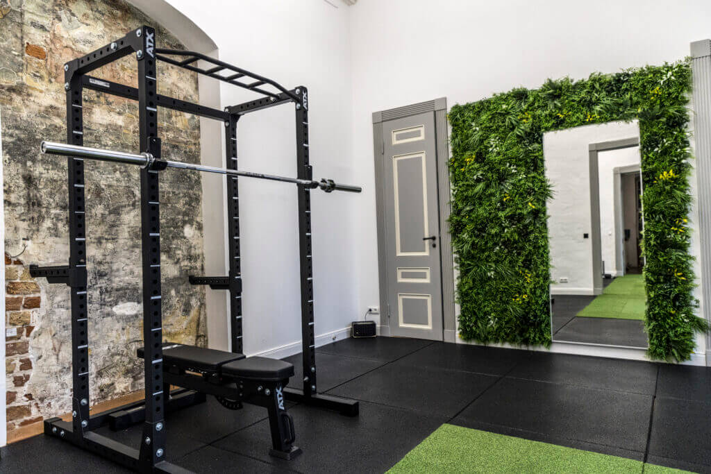 Ein moderner Fitnessraum des Personal Training Studios Lübeck mit einem Squat Rack und einer Bank, schwarzem Gummiboden, freiliegender Backsteinwand, großem Spiegel und einem üppigen grünen vertikalen Garten neben dem Spiegel.