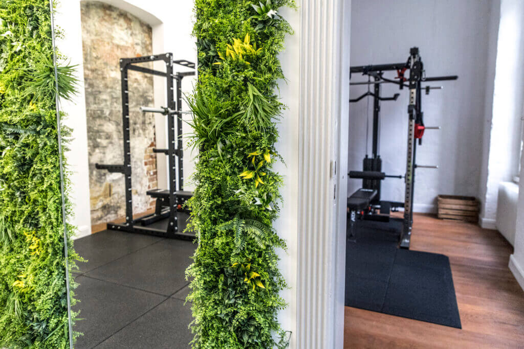 Ein moderner Fitnessraum mit Krafttrainingsgeräten, sichtbar durch eine Türöffnung. Im Vordergrund befindet sich eine Wand mit üppigen Grünpflanzen, die dem Fitnessraum einen Hauch von Natur verleihen.