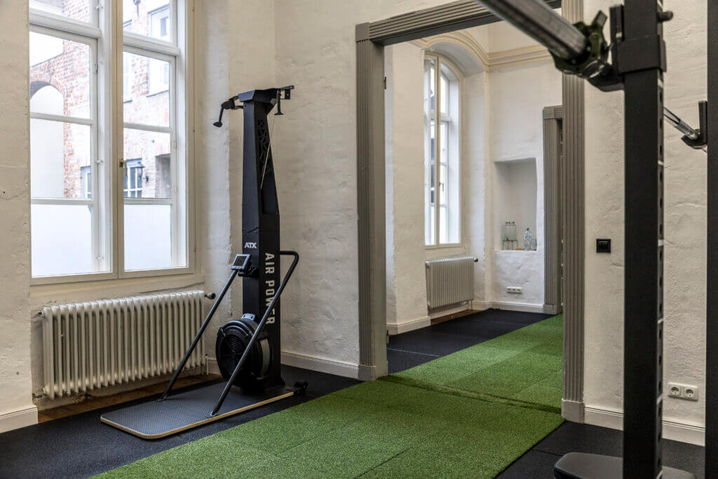 Ein helles, modernes Personal Training Studio Lübeck mit weißen Wänden, großen Fenstern, grünem Rasenboden, einem SkiErg-Gerät und einem Regal mit Wasserflaschen im Hintergrund.