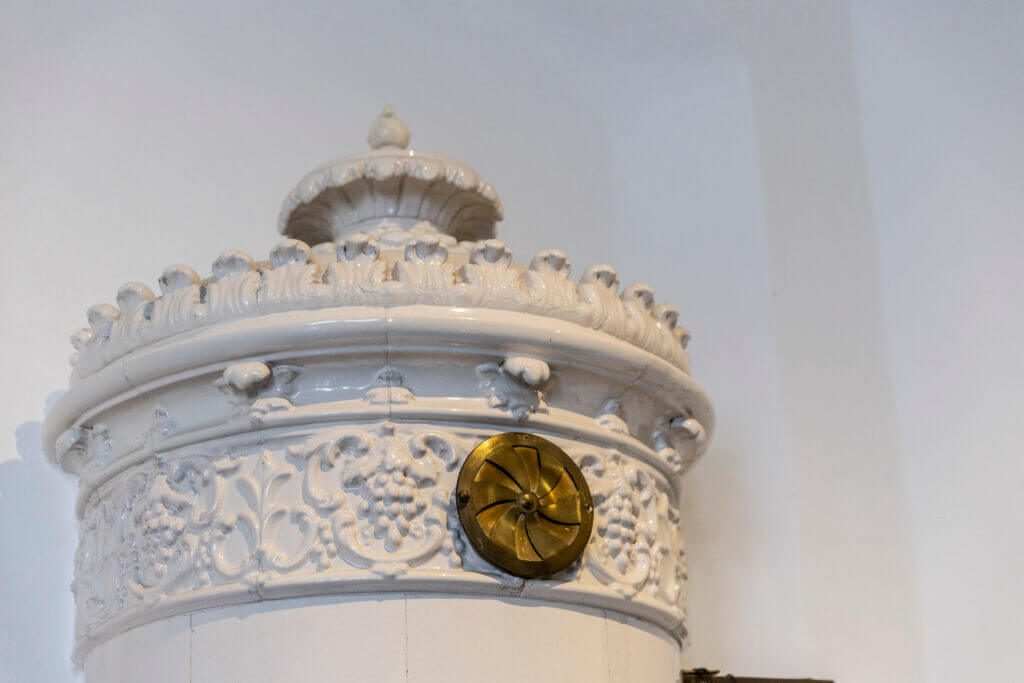 Verschnörkelter weißer Keramikofen mit dekorativen Blumenmustern und einer Lüftungsabdeckung aus Messing, der einem Personal Training Studio Lübeck vor einer schlichten weißen Wand Charme verleiht.