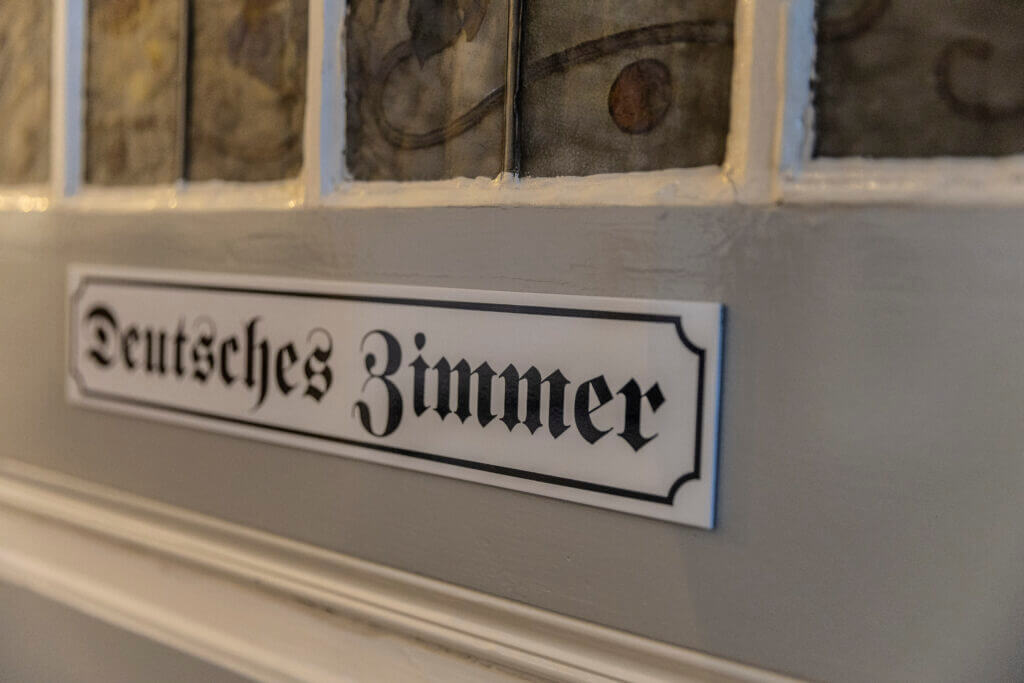 Ein weißes Schild mit schwarzer gotischer Schrift mit der Aufschrift Deutsches Zimmer ist an einer grauen Wand unter dekorativen Glasscheiben angebracht und erinnert an die klassische Beschilderung eines Personal Training Studios in Lübeck.