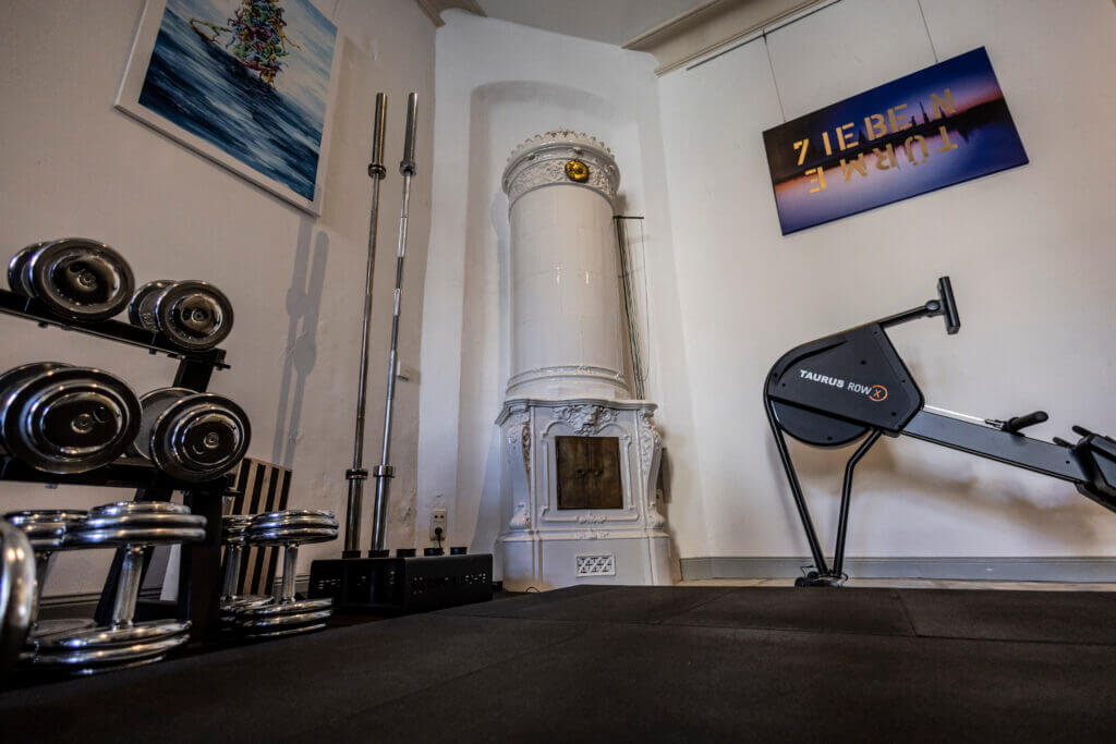 Ein Personal Training Studio in Lübeck mit Hanteln, Langhanteln, einem Rudergerät und einem weißen, dekorativen Keramikofen. Gemälde und an der Wand befestigte Spiegel oder Kunstwerke schmücken die Wände, während schwarze Matten den Boden bedecken.