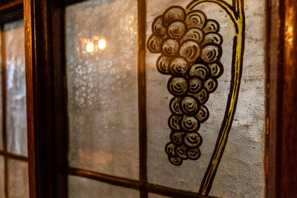 Ein Buntglasfenster mit einer Traube aus dunkelvioletten Trauben mit gelbem Stiel vor einem strukturierten, leicht nebligen Hintergrund - wie das einladende Ambiente eines Personal Training Studios in Lübeck. Das warme Innenlicht spiegelt sich schwach im Glas.