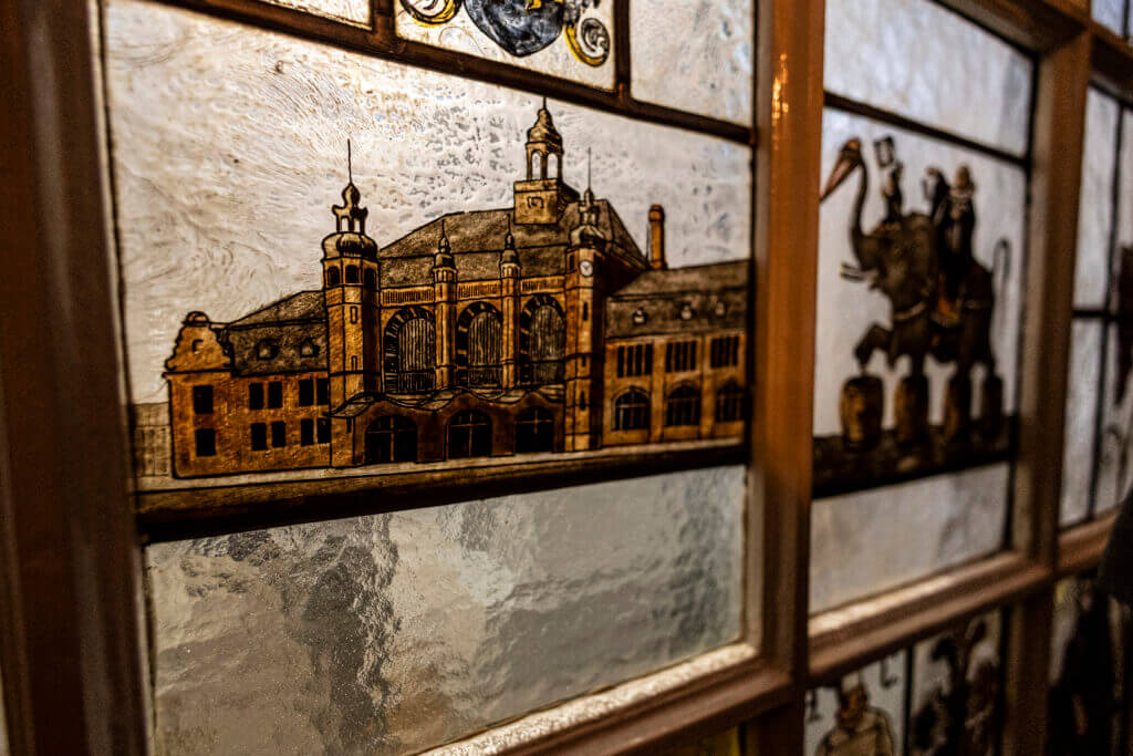 Buntglasfenster mit einer detaillierten Abbildung eines historischen Gebäudes mit Türmen und Bogenfenstern, eingefasst in einen Holzrahmen. Das Vintage-Glasdesign spiegelt den zeitlosen Charme des Personal Training Studios Lübeck wider.