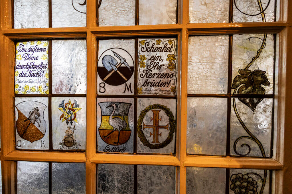 Buntglasfenster mit verschiedenen gemalten Tafeln mit deutschem Text, Symbolen wie einem Kreuz und einem Stern, einem Vogel, Werkzeugen und verschnörkelten Blättern - ähnlich der Kunst im Personal Training Studio Lübeck. Das Glas hat einen gelblichen Rahmen und strukturierte Abschnitte.