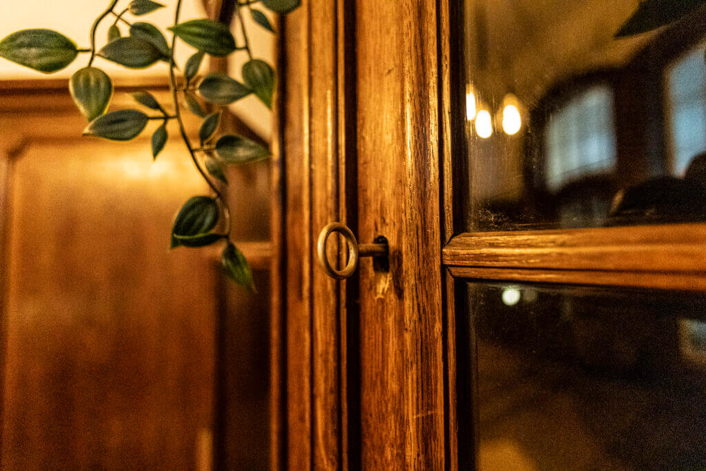 Eine Nahaufnahme einer hölzernen Schranktür mit einem runden Metallschlüssel im Schloss, Glasscheiben und einer grünen Blattpflanze in der Nähe in einem Personal Training Studio in Lübeck. Warmes Licht wird von den Glasscheiben reflektiert und schafft eine gemütliche Atmosphäre.