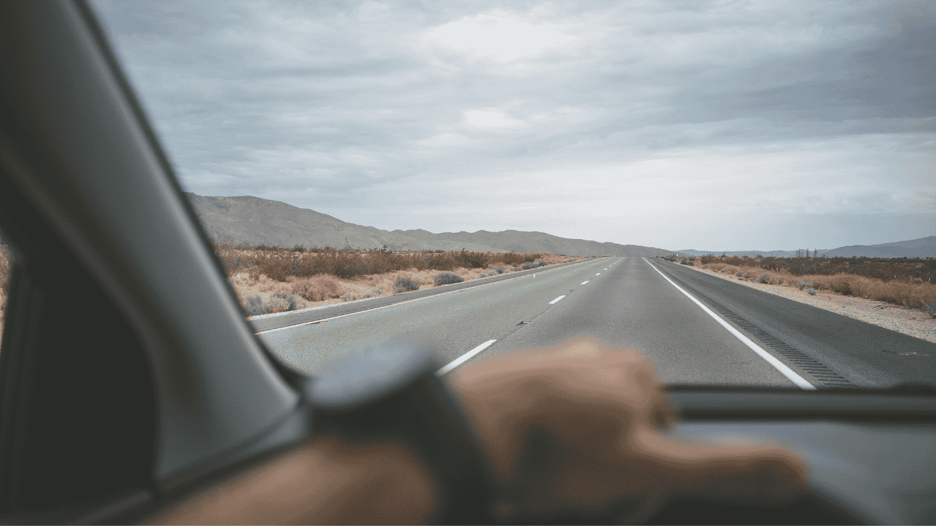 Blick aus dem Inneren eines Autos auf eine Person am Lenkrad, die eine leere Wüstenstraße unter bewölktem Himmel entlang fährt, mit Bergen in der Ferne.